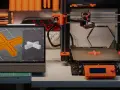 Na stole warsztatowym widać różne rodzaje drukarek 3D, tablet z modelem 3D i wydrukowany element.