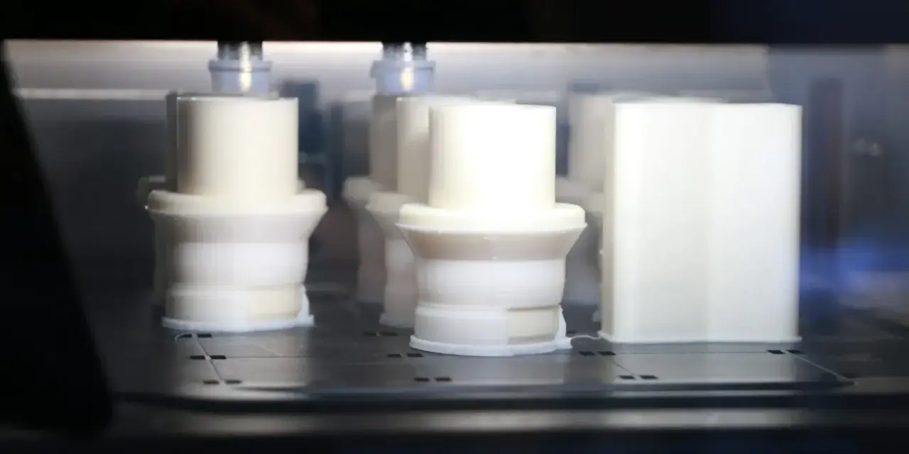 Białe obiekty w drukarce 3D, z widocznym elementem przypominającym 3d print raft, tworzą się warstwa po warstwie.
