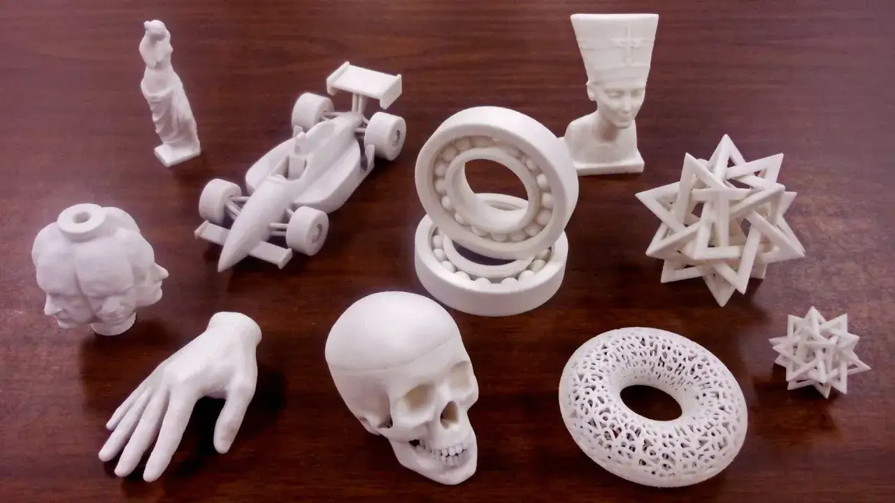 R&oacute;żnorodne modele wydrukowane na drukarce 3d: czaszka, dłoń, bolid F1, łożyska, popiersie Nefertiti, wielogłowa postać, figurka i geometryczne bryły.